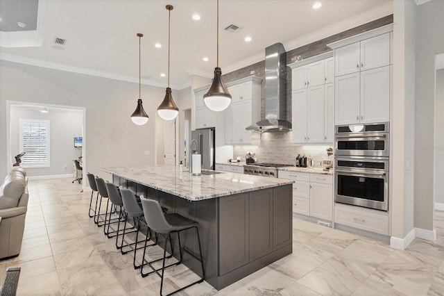 kitchen featuring a large island with sink, wall chimney range hood, appliances with stainless steel finishes, decorative light fixtures, and light stone counters