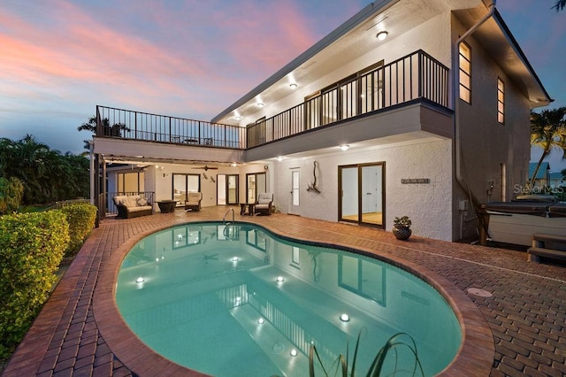 back house at dusk with a balcony, a patio area, a swimming pool with hot tub, ceiling fan, and an outdoor living space