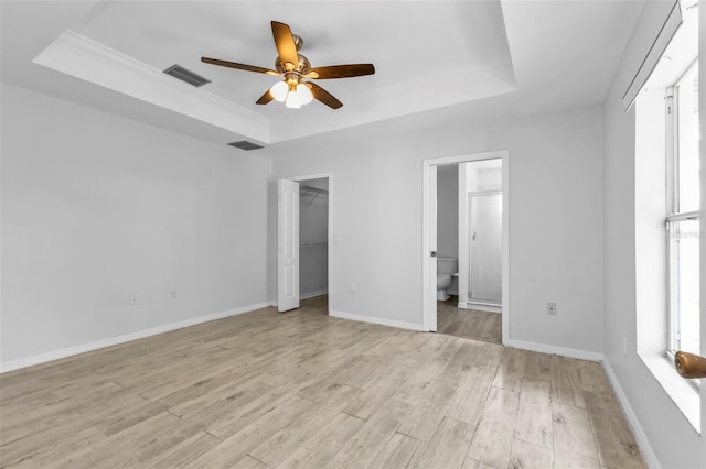 unfurnished bedroom with a raised ceiling, connected bathroom, ceiling fan, light wood-type flooring, and a walk in closet