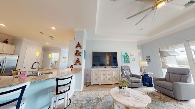 living room with a raised ceiling, light hardwood / wood-style floors, ornamental molding, ceiling fan, and sink