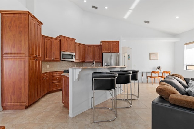 kitchen with a kitchen bar, stainless steel appliances, backsplash, high vaulted ceiling, and a center island with sink