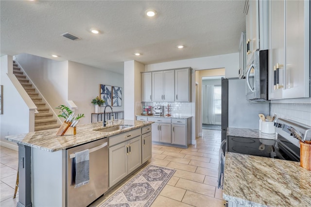 kitchen featuring sink, a center island with sink, appliances with stainless steel finishes, gray cabinets, and light stone countertops