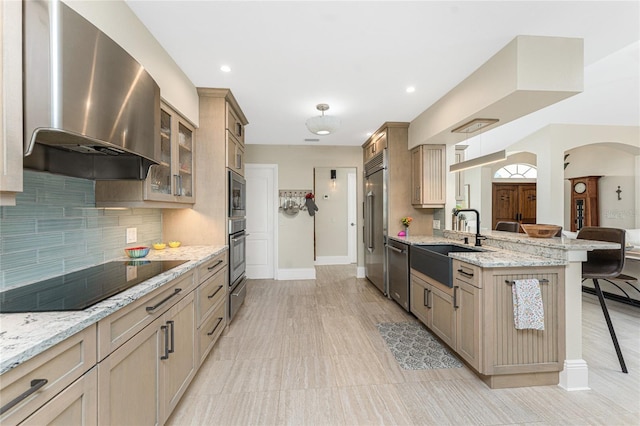 kitchen featuring a kitchen bar, sink, light stone counters, built in appliances, and range hood