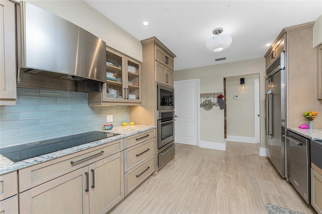 kitchen featuring light stone counters, stainless steel appliances, light brown cabinetry, and wall chimney range hood