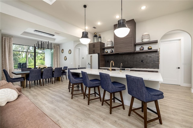 kitchen with sink, hanging light fixtures, a kitchen island with sink, light wood-type flooring, and high end white refrigerator