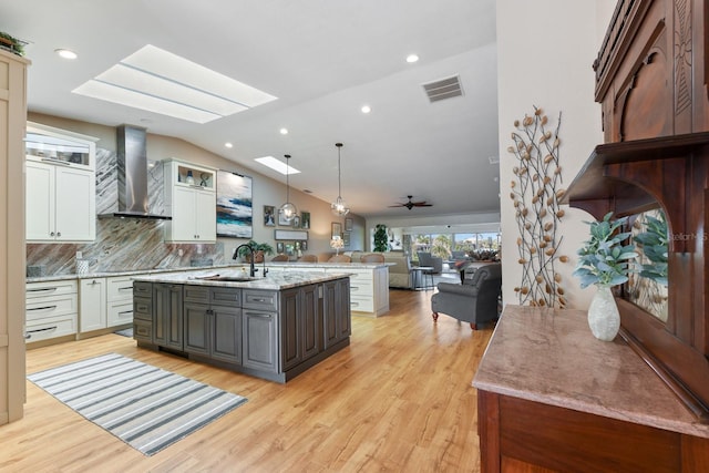 kitchen with sink, white cabinetry, pendant lighting, a kitchen island with sink, and wall chimney range hood