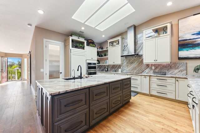 kitchen featuring a skylight, an island with sink, sink, light stone countertops, and wall chimney range hood