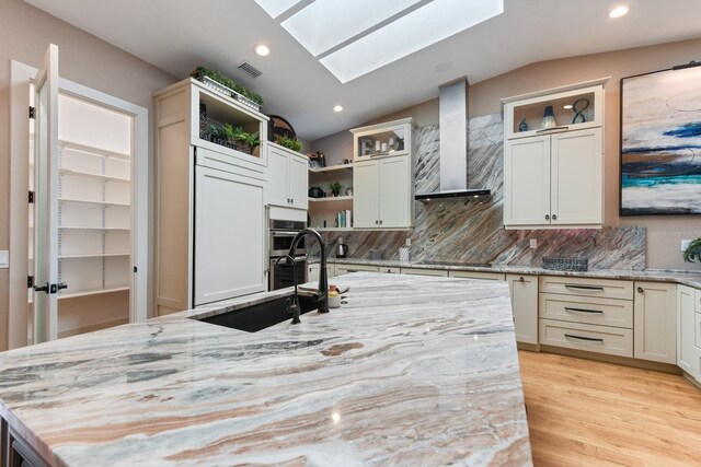 kitchen with sink, a kitchen island with sink, light stone counters, paneled fridge, and wall chimney exhaust hood