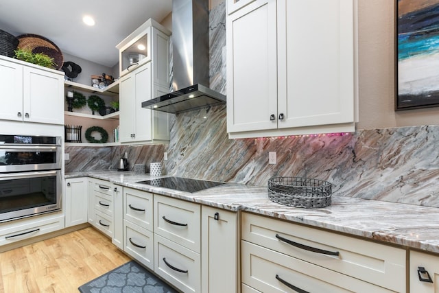 kitchen featuring double oven, backsplash, light stone counters, black electric cooktop, and wall chimney exhaust hood