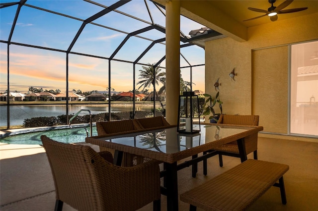 patio terrace at dusk featuring a water view, ceiling fan, and glass enclosure