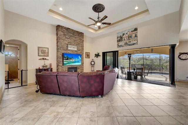 living room featuring a high ceiling, a stone fireplace, ceiling fan, and a tray ceiling