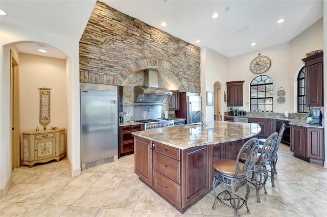 kitchen with stainless steel built in refrigerator, an island with sink, a kitchen breakfast bar, light stone countertops, and wall chimney exhaust hood