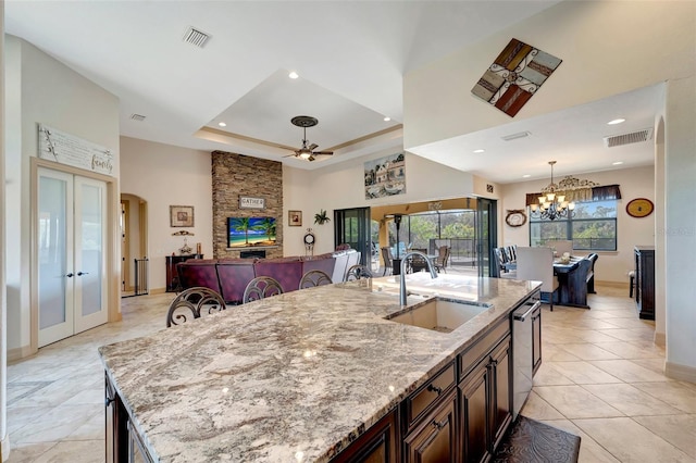 kitchen with sink, light stone counters, a center island with sink, stainless steel dishwasher, and a raised ceiling