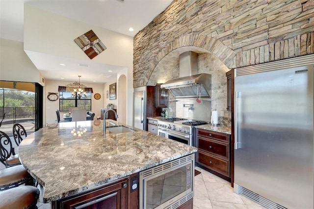 kitchen featuring sink, a kitchen island with sink, built in appliances, light stone counters, and wall chimney exhaust hood