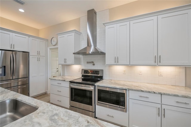 kitchen featuring wall chimney exhaust hood, white cabinetry, appliances with stainless steel finishes, light stone countertops, and backsplash