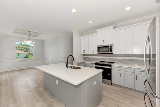 kitchen with sink, a center island with sink, ornamental molding, appliances with stainless steel finishes, and a tray ceiling