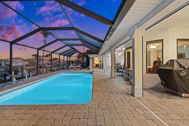 pool at dusk featuring a patio area, grilling area, a water view, and glass enclosure