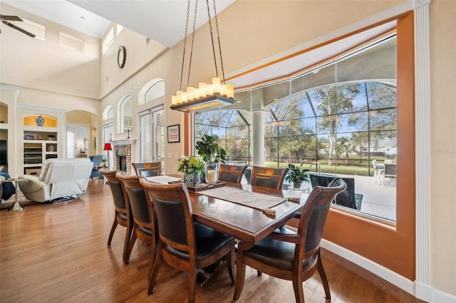 dining area featuring a high ceiling, wood-type flooring, a brick fireplace, and ceiling fan with notable chandelier