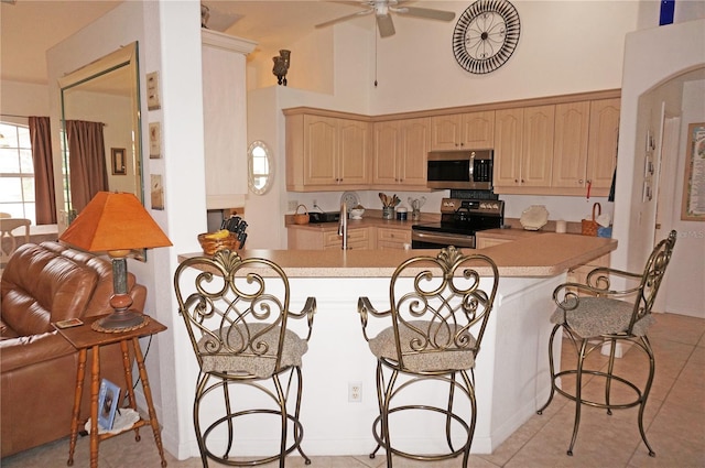 kitchen with light tile patterned floors, appliances with stainless steel finishes, a kitchen bar, kitchen peninsula, and light brown cabinets