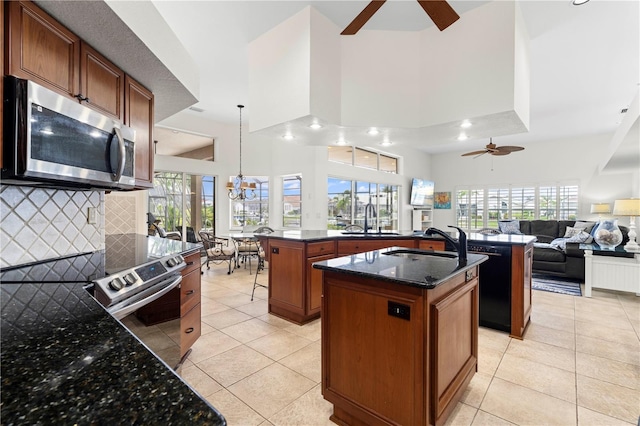 kitchen featuring dark stone countertops, hanging light fixtures, stainless steel appliances, a center island with sink, and ceiling fan with notable chandelier