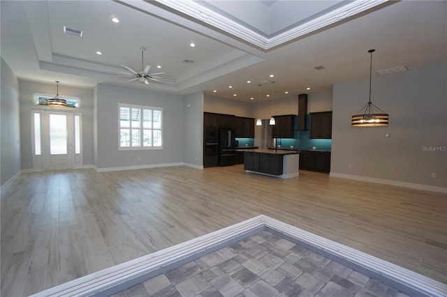 kitchen featuring wall chimney range hood, a tray ceiling, light hardwood / wood-style floors, and an island with sink