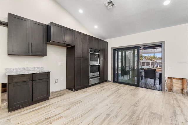 kitchen with lofted ceiling, dark brown cabinets, light wood-type flooring, double oven, and light stone countertops