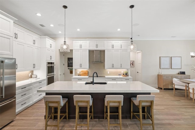 kitchen with stainless steel appliances, a kitchen island with sink, wall chimney range hood, and decorative light fixtures