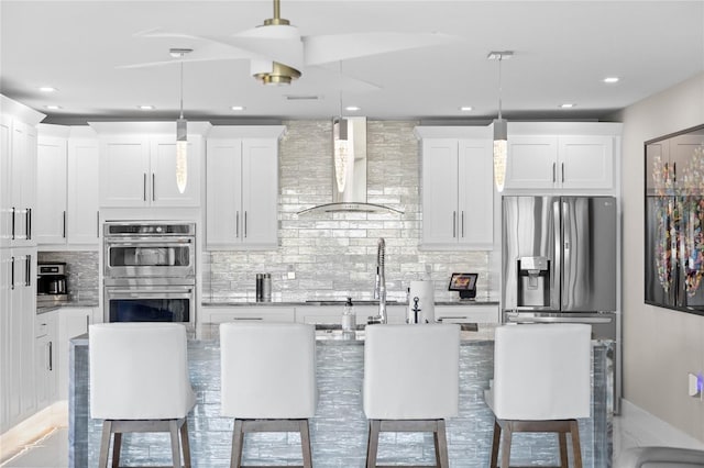 kitchen featuring pendant lighting, white cabinetry, an island with sink, and wall chimney range hood