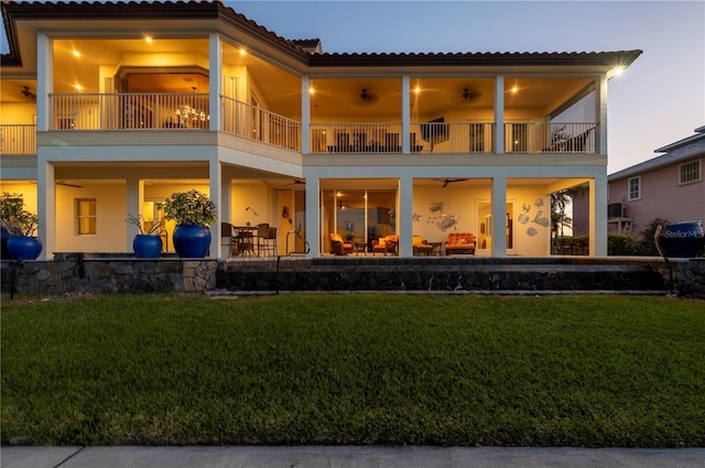 back house at dusk with a balcony, a patio, and a lawn