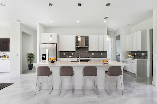 kitchen with stainless steel refrigerator with ice dispenser, wall chimney exhaust hood, sink, white cabinetry, and hanging light fixtures