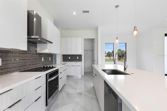 kitchen with appliances with stainless steel finishes, sink, white cabinets, hanging light fixtures, and wall chimney range hood