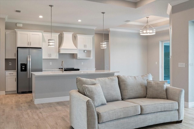 kitchen featuring stainless steel refrigerator with ice dispenser, premium range hood, an island with sink, and hanging light fixtures