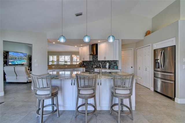 kitchen with stainless steel refrigerator with ice dispenser, wall chimney exhaust hood, decorative light fixtures, light stone countertops, and white cabinets