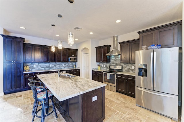 kitchen with sink, hanging light fixtures, a center island with sink, appliances with stainless steel finishes, and wall chimney range hood