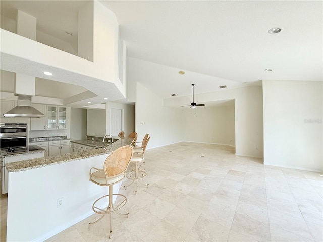 kitchen with wall chimney range hood, ceiling fan, double oven, light stone countertops, and kitchen peninsula