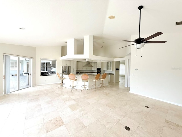 kitchen featuring stainless steel refrigerator with ice dispenser, high vaulted ceiling, a kitchen breakfast bar, ceiling fan, and wall chimney range hood