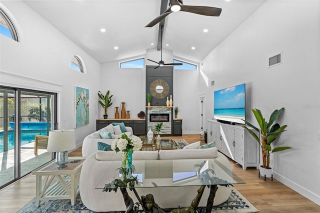 living area with visible vents, light wood-style floors, a glass covered fireplace, high vaulted ceiling, and baseboards