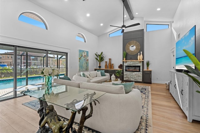 living area featuring high vaulted ceiling, a stone fireplace, light wood-style flooring, a ceiling fan, and beam ceiling