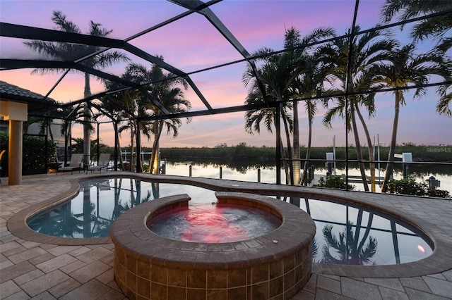 pool at dusk featuring an in ground hot tub, a water view, glass enclosure, and a patio