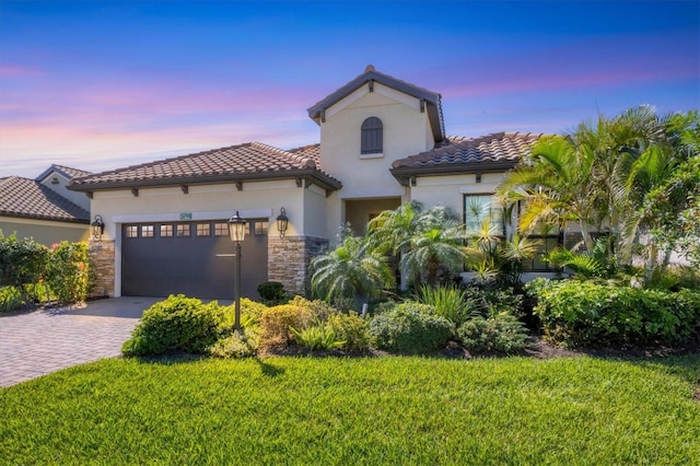 mediterranean / spanish-style home featuring stone siding, an attached garage, decorative driveway, a front yard, and stucco siding