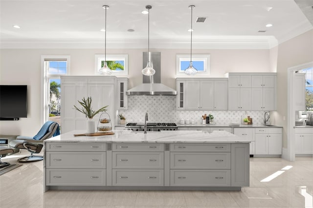 kitchen featuring gray cabinetry, wall chimney range hood, a kitchen island with sink, and hanging light fixtures