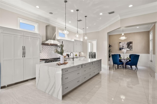 kitchen featuring hanging light fixtures, an island with sink, wall chimney range hood, and light stone counters