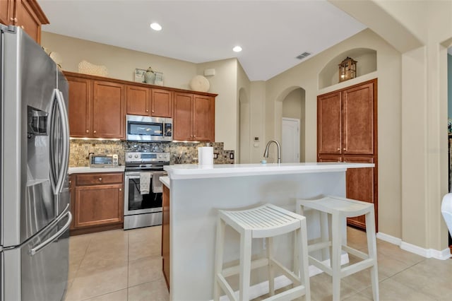 kitchen with light tile patterned flooring, appliances with stainless steel finishes, a breakfast bar, decorative backsplash, and a kitchen island with sink