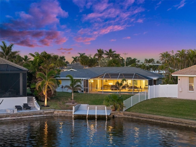 back house at dusk featuring a water view, glass enclosure, and a lawn