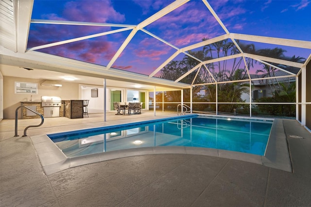 pool at dusk with exterior kitchen, grilling area, glass enclosure, and a patio area