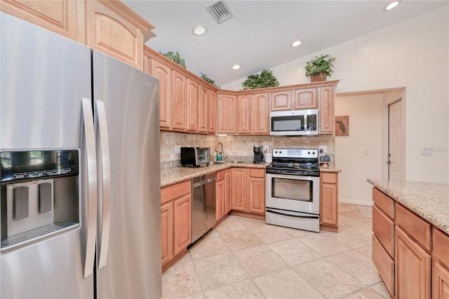 kitchen with appliances with stainless steel finishes, lofted ceiling, backsplash, light stone countertops, and light brown cabinets