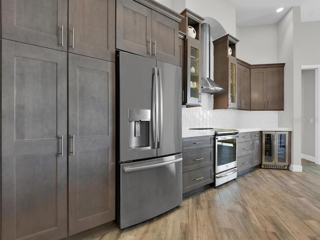 kitchen with wall chimney range hood, light hardwood / wood-style flooring, appliances with stainless steel finishes, tasteful backsplash, and beverage cooler