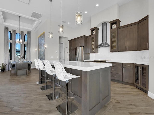 kitchen with wine cooler, wall chimney exhaust hood, hanging light fixtures, stainless steel appliances, and a kitchen island with sink