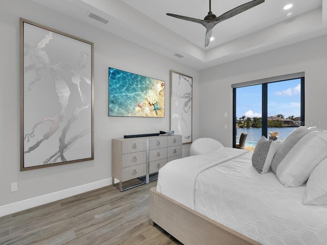 bedroom with access to exterior, ceiling fan, light hardwood / wood-style floors, a raised ceiling, and a water view