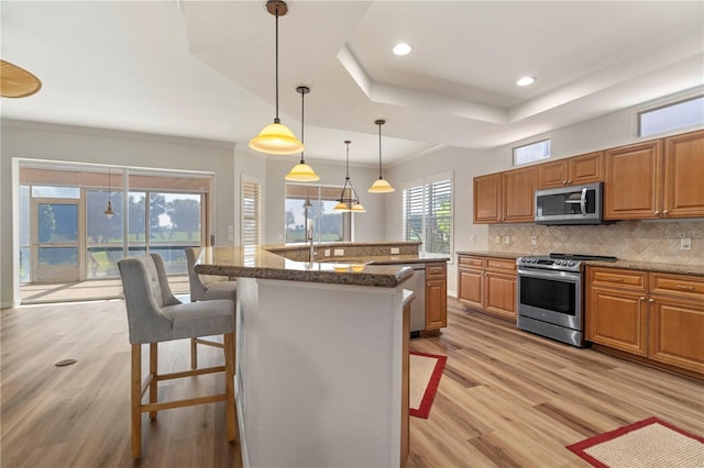 kitchen with a breakfast bar area, a kitchen island with sink, stainless steel appliances, brown cabinetry, and a tray ceiling